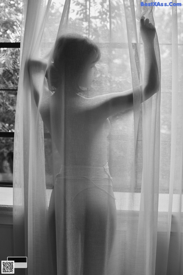 A woman standing in front of a window with sheer curtains.