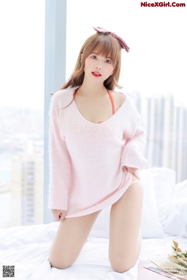 A woman in a pink sweater sitting on a bed.